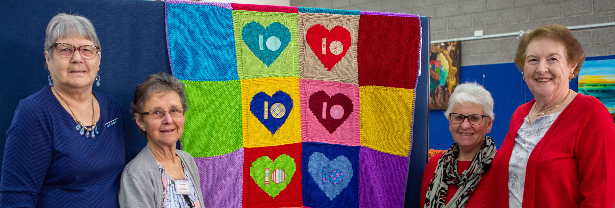 Wrap with Love knitters Jan Riddell, Loelene Harrison, Ann French and Sandra Brooks celebrate the groups 10 year anniversary. Four knitters standing with a rug