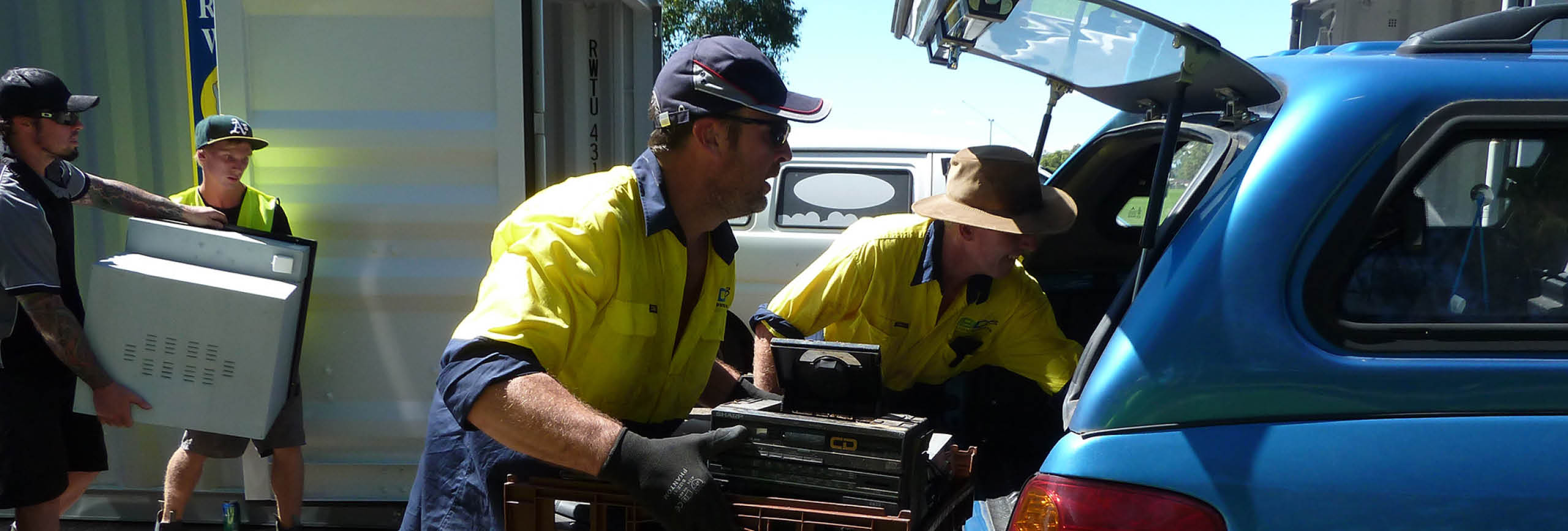 Workmen in fluorescent tops loading electronic waste into a car, one giving thumbs up