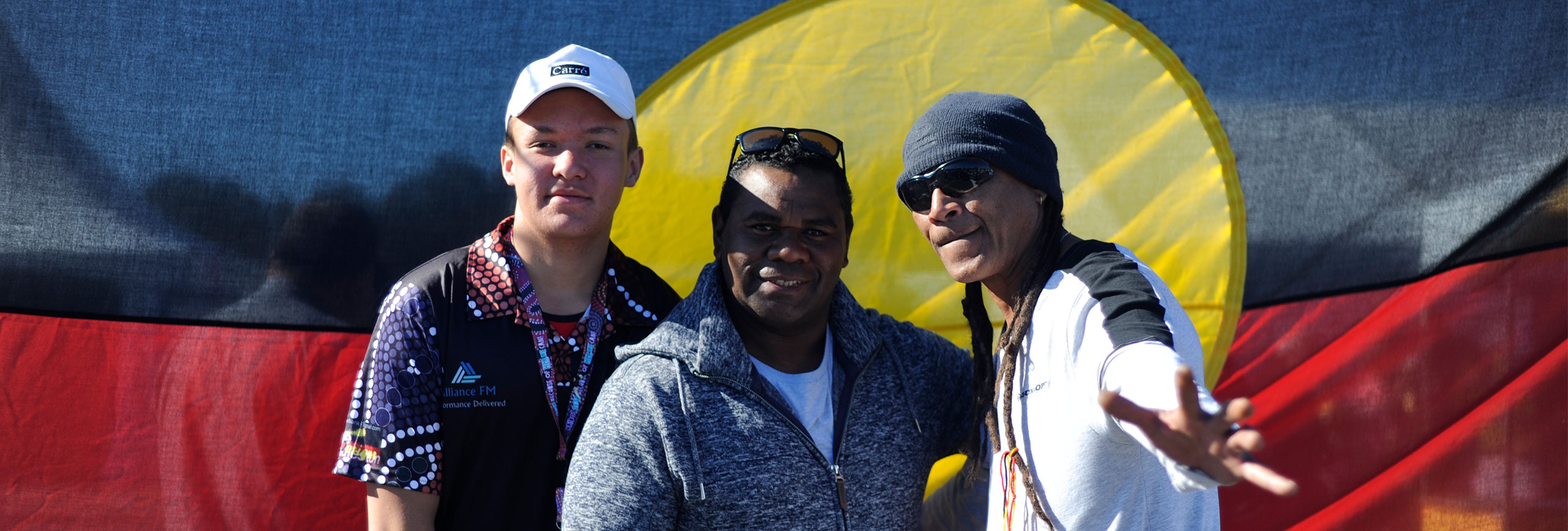 Three indigenous men standing in front of Aboriginal flag