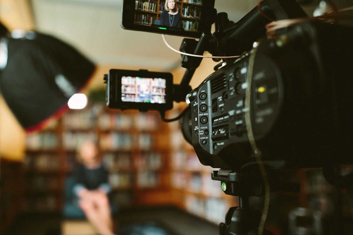 video camera in foreground, pointing at person sitting on chair in distance