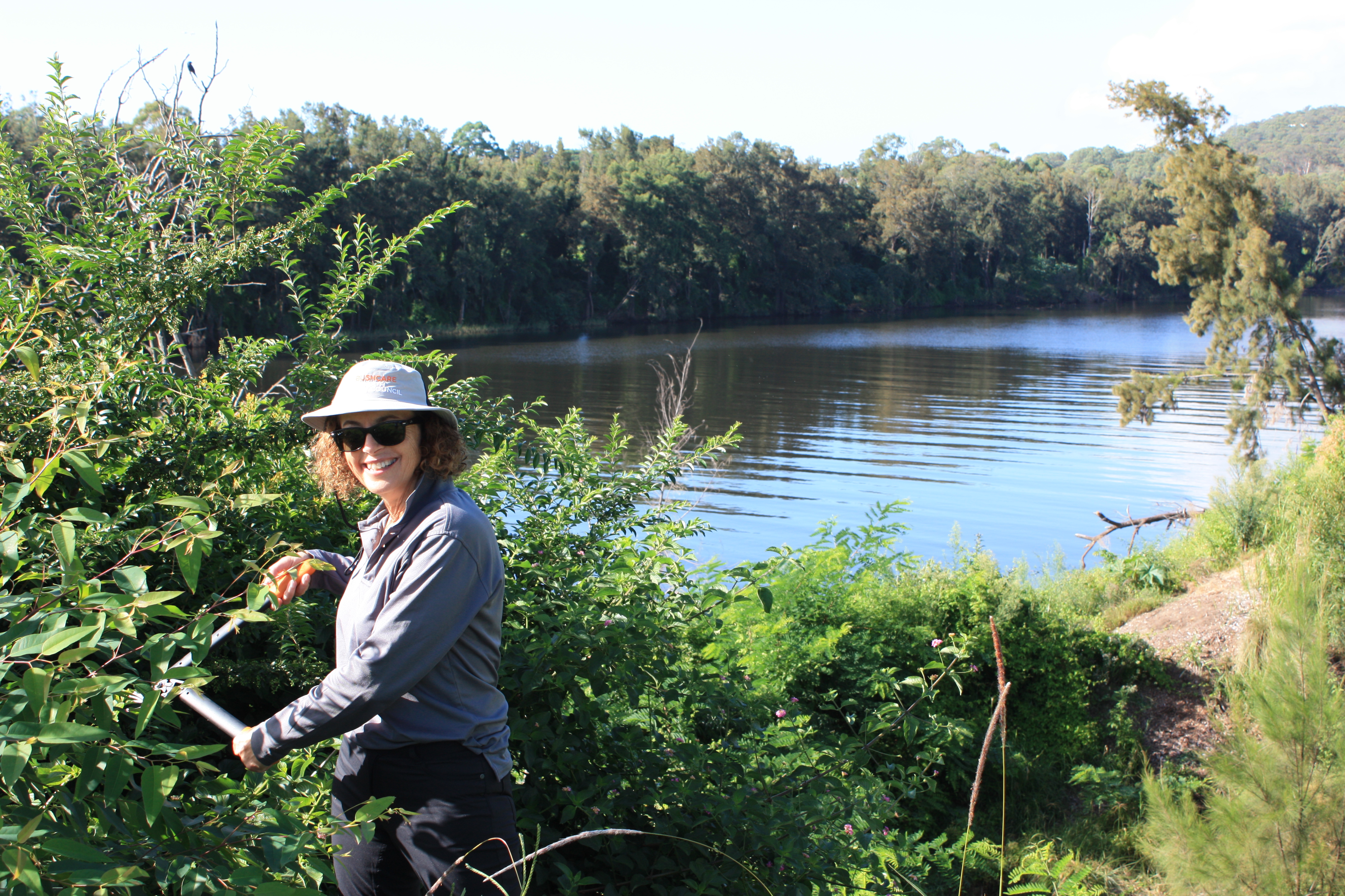 A volunteer smiling as they remove weeds from plants along the riverbank.